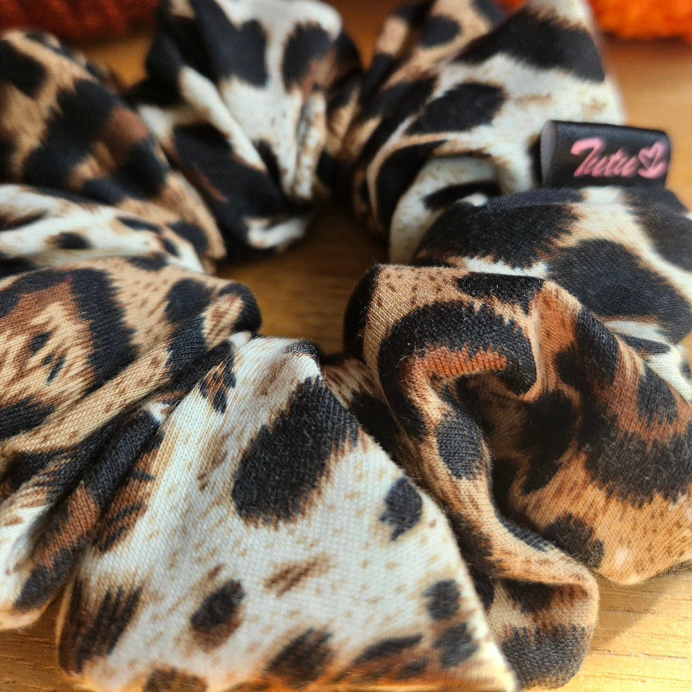 A leopard print fabric scrunchie with a small black brand tag reading 'Tutu22', displayed on a wooden surface with decorative orange pumpkins in the background.