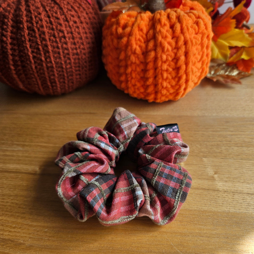 A fabric scrunchie with a orange, brown, and beige plaid pattern sits on a wooden table, styled with autumn-themed crochet pumpkins in the background.