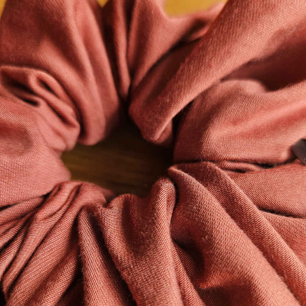 A close-up of a burnt orange or terracotta scrunchie made of stretch bamboo and elastane fabric, shown against a light wood background.