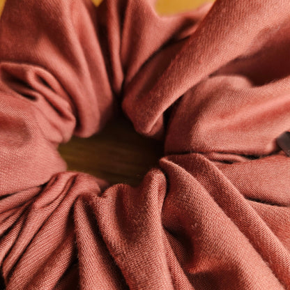 A close-up of a burnt orange or terracotta scrunchie made of stretch bamboo and elastane fabric, shown against a light wood background.
