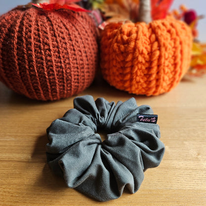 A olive green fabric scrunchie with a small black 'Tutu22' brand tag, displayed on a wooden table with decorative knitted pumpkins in the background.