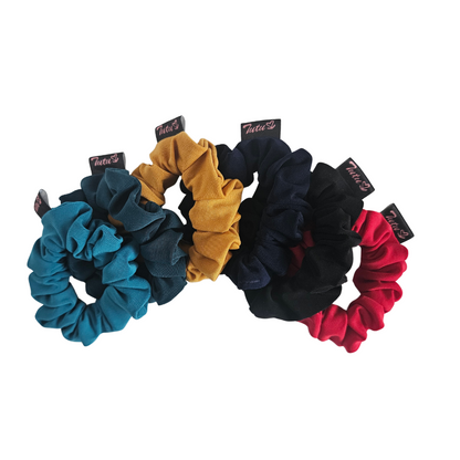 Six back to school fabric hair scrunchies in assorted colours (teal, bottle green, mustard, navy, black, and red) arranged in a fan shape on a plain white background. Each scrunchie has a small black fabric tag with the brand name 'Tutu22' printed on it.