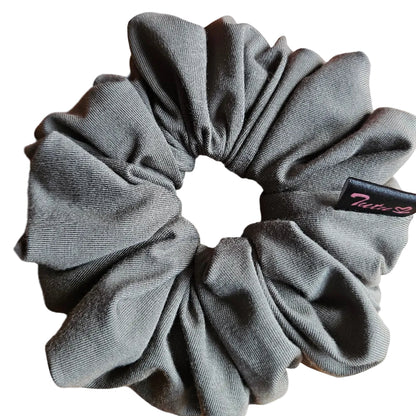 A olive green fabric scrunchie with a small black brand tag, resting on a light brown wooden surface.