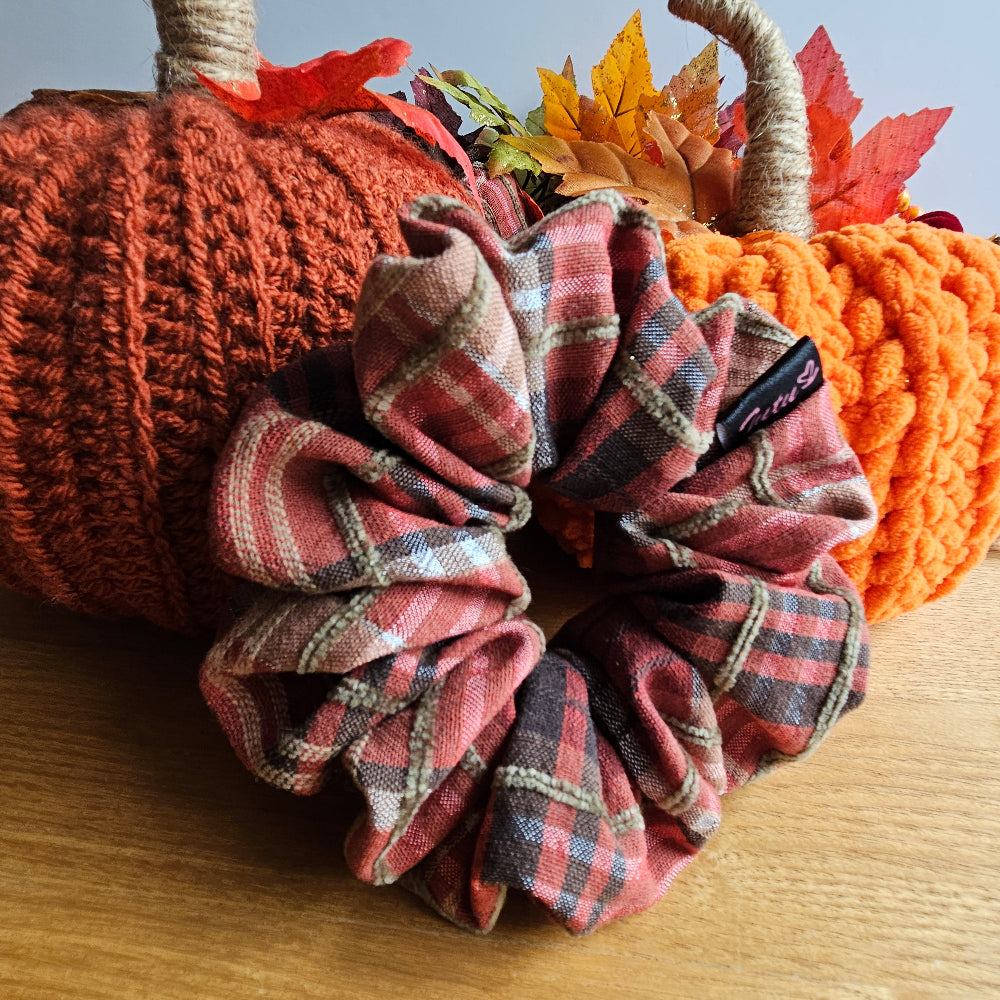A fabric scrunchie in a plaid pattern, styled in front of two decorative crochet pumpkins on a wooden surface.