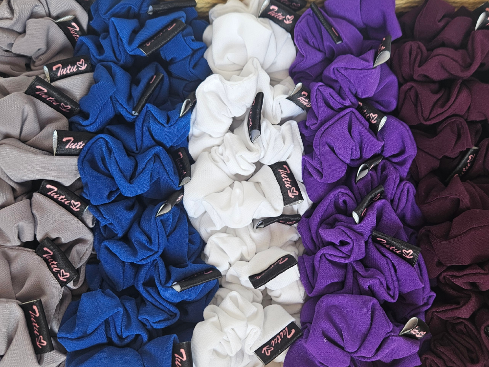 A close-up arrangement of fabric hair scrunchies in multiple colors, including gray, royal blue, white, purple, and maroon, each with a small black tag that reads 'Tula's'.