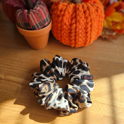 A leopard print fabric scrunchie with a small black brand tag, displayed on a wooden table with autumn-themed decorative pumpkins in the background.