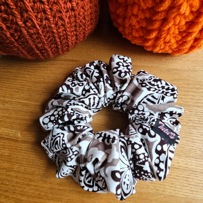 A fabric hair scrunchie with a black and white floral pattern sits on a wooden tabletop. In the background, partially cropped, are two crochet pumpkins in rust orange and bright orange.